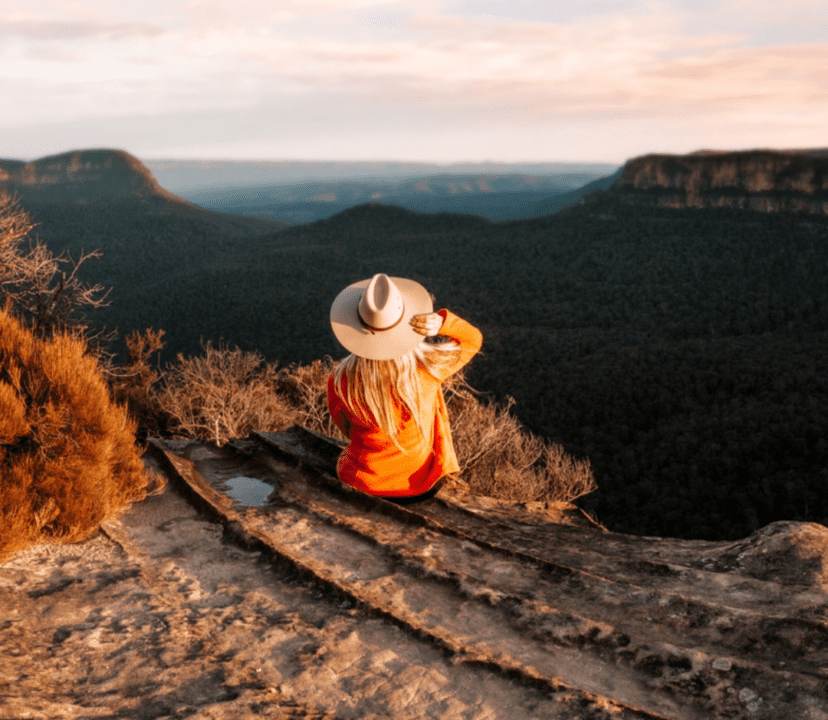 Solo female traveller sat on top of a mountain looking out at the sunset holding her hat embracing her wanderlust.