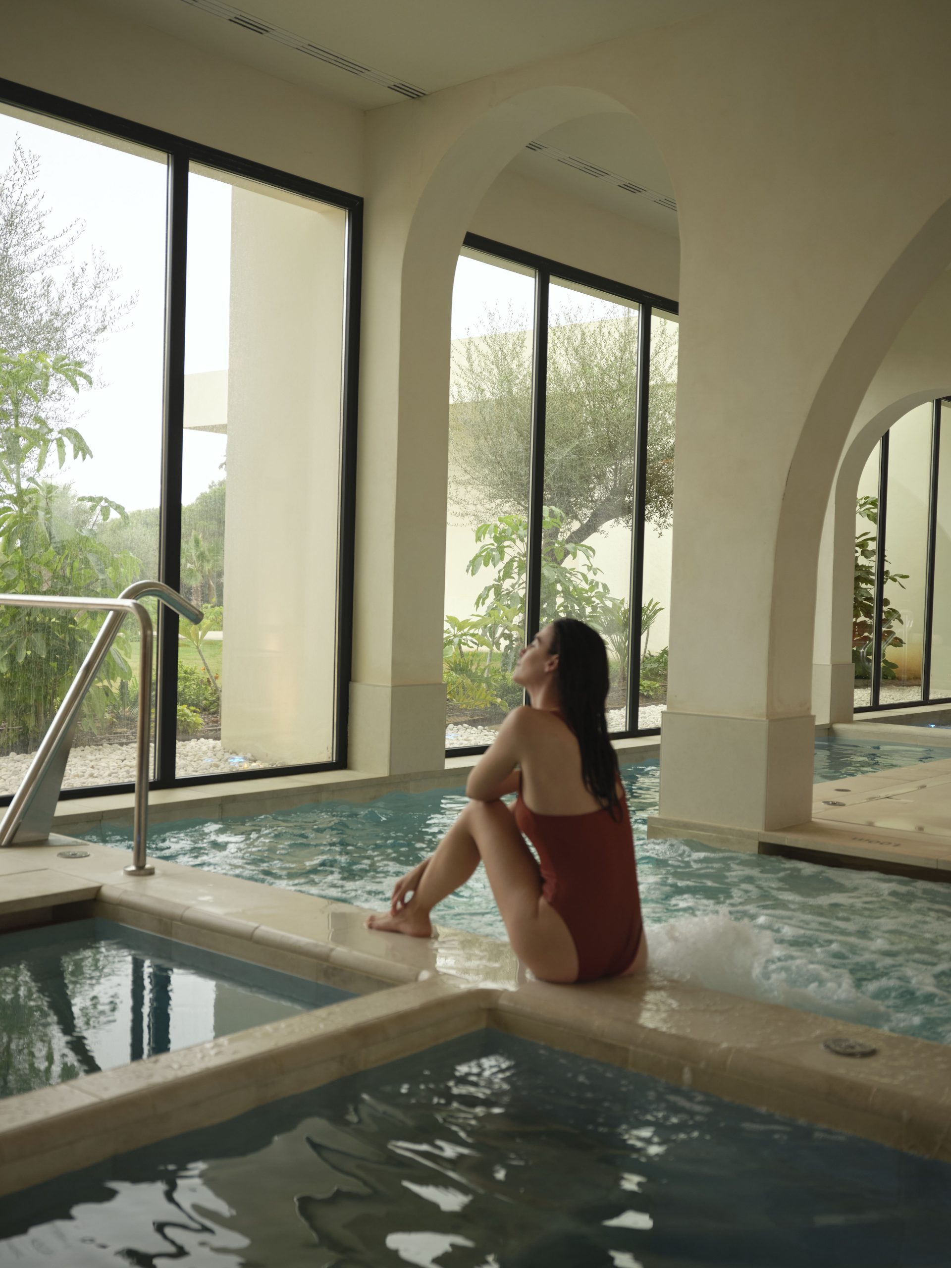 A woman sat gracefully on the edge of the indoor pool at the luxury hotel whilst looking out of the large windows that let in natural light.