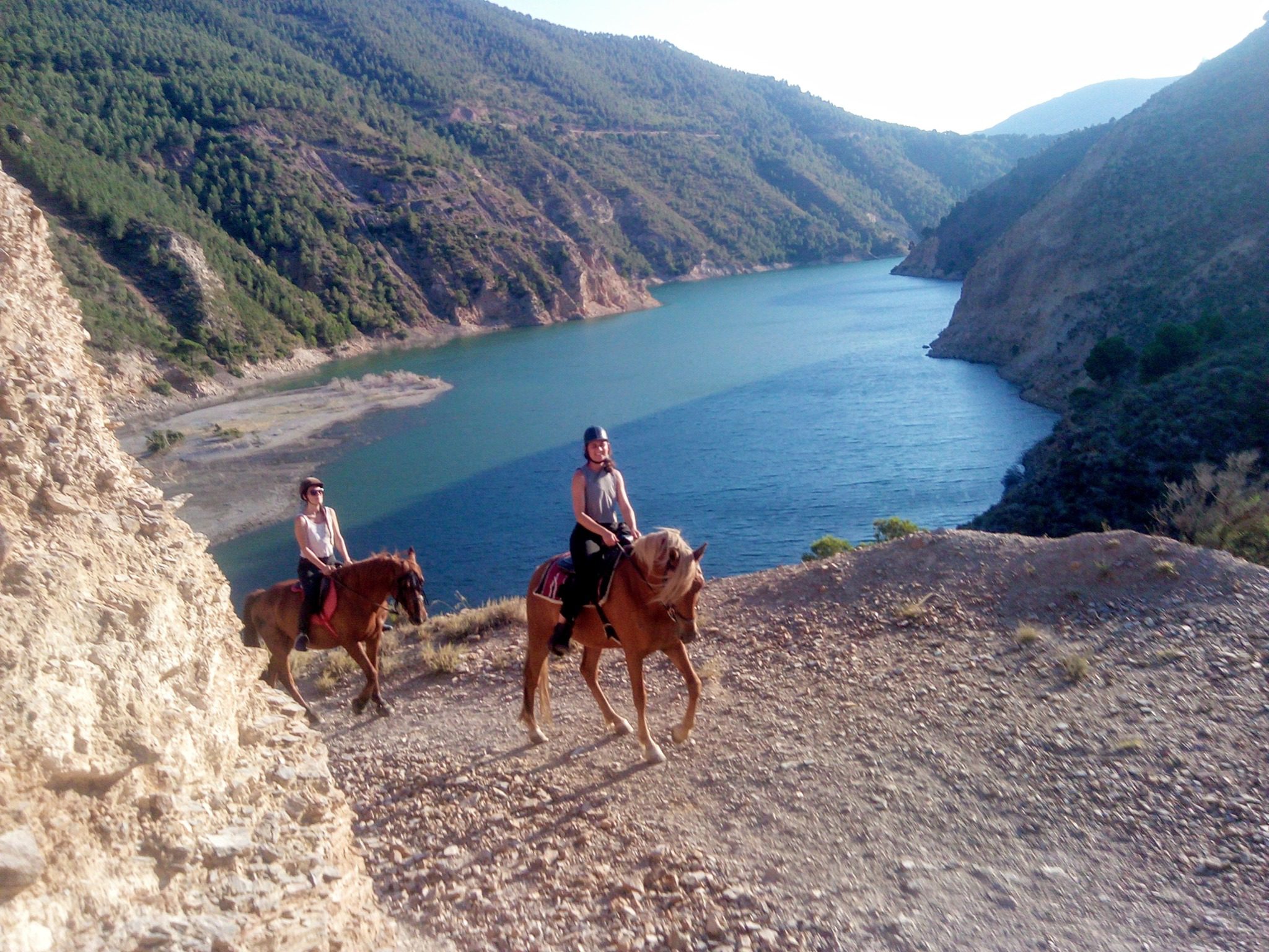 2 horseback riders on a 3 hour trek coming towards the camera with a reservoir and green mountainside behind in Granada Spain