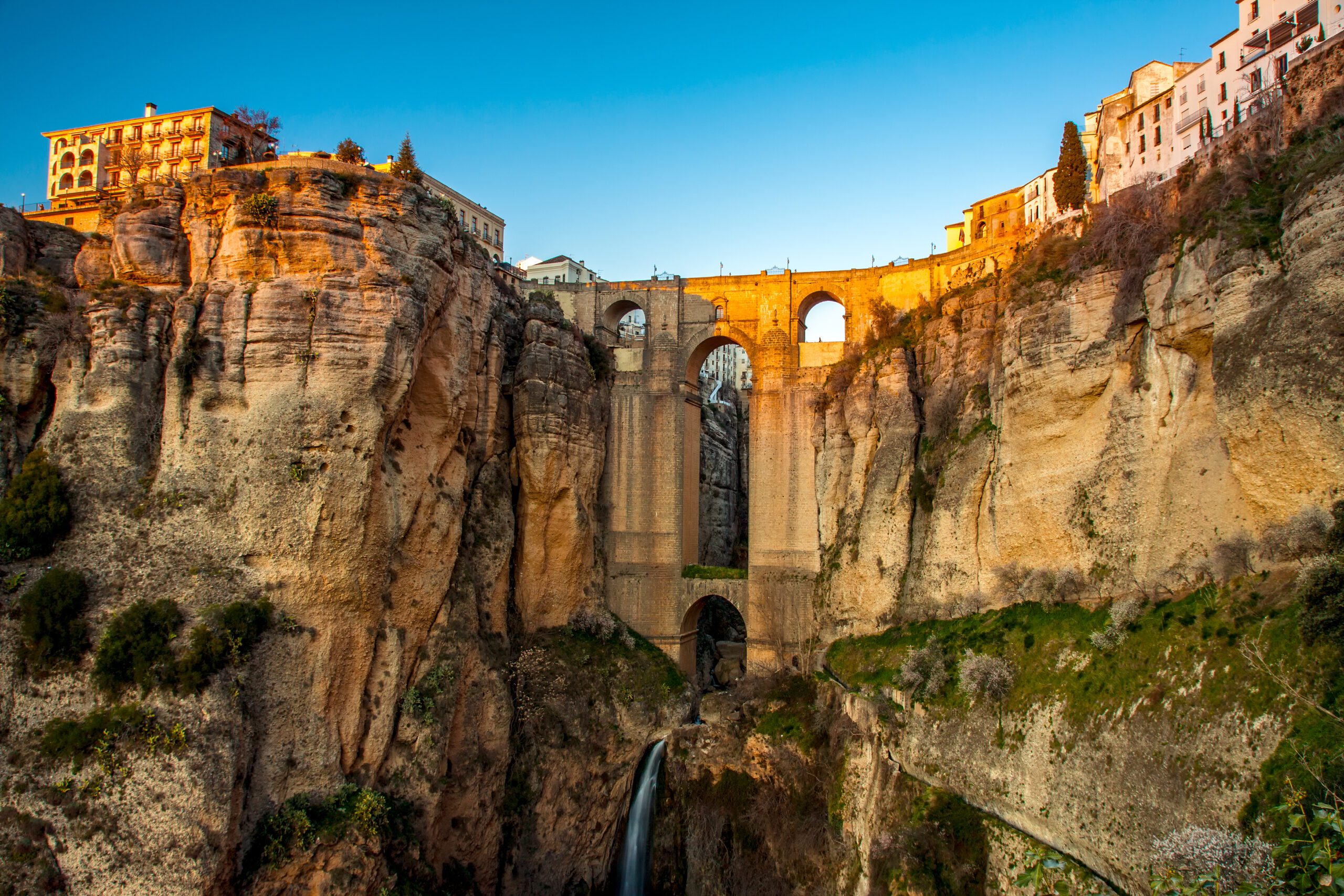 The famous bridge that enables access to the ancient city of Ronda showing the water running through teh stream at the bottom of the valley and the sun setting on the bridge with deep blue skies.