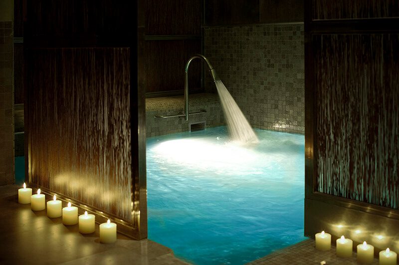 An indoor pool at the luxury spa with a water feature flowing into the water and candles lit around the edge of the pool.