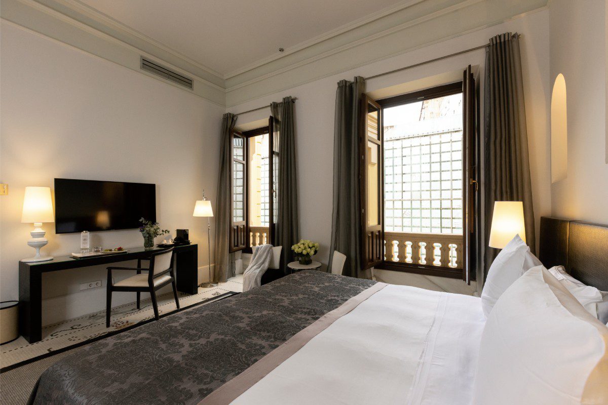 A bedroom at the luxury hotel with a large bed, a desk and chair, a TV and two double windows.