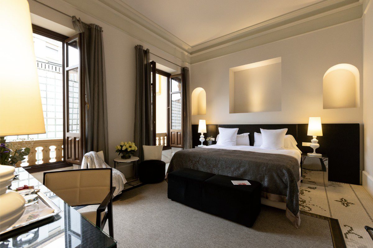 A bedroom in the luxury hotel with a large bed, a desk, two chairs next to a small table and two double windows.