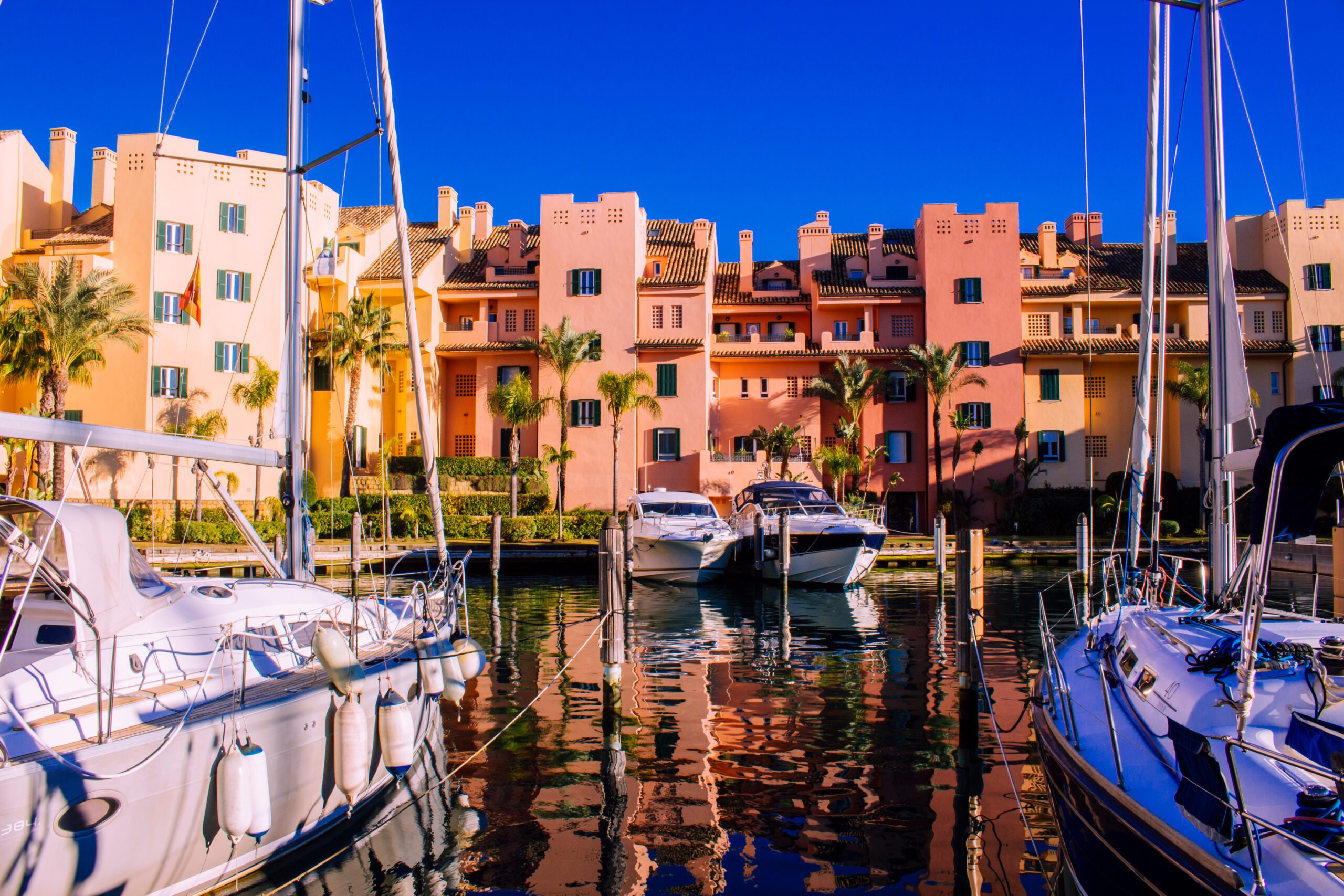 Luxury yachts berthed in the canals of Sotogrande Marina with the apartments painted in the Andalucian reds, creams and yellows behind under a deep blue sky.