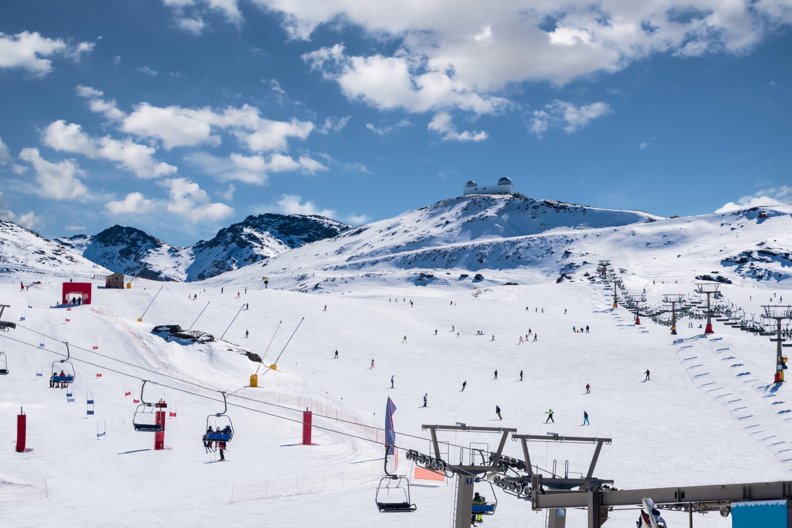 Snowy ski slopes with skiiers and the ski lifts with mountain tops behind and blue sky with white fluffy clouds in the Sierra Nevada in Granada Spain