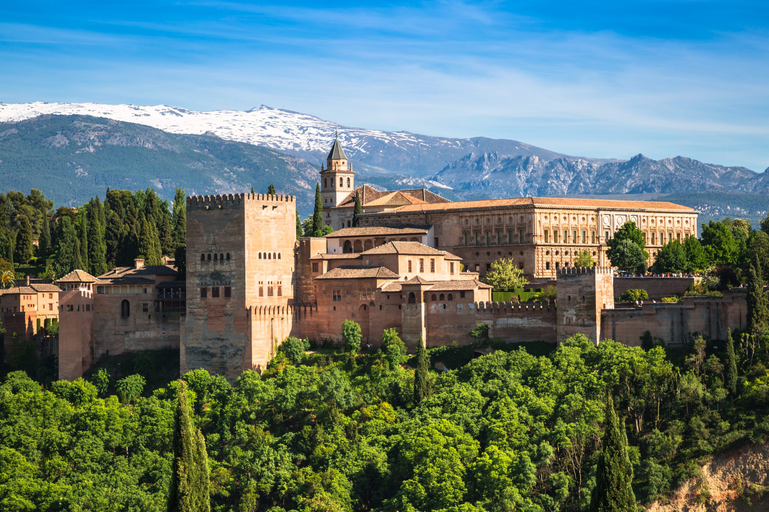 A view of Alhambra, Granada surrounded by greenery with mountains and a blue sky behind.