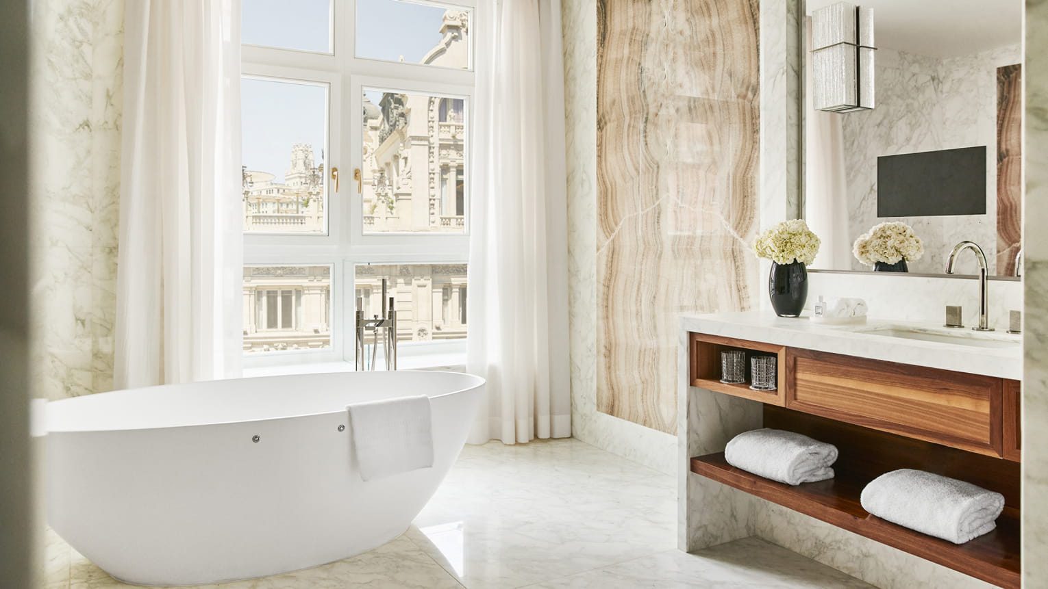 A luxury bathroom featuring a large free standing bath in the centre and a large window, showcasing the views of Madrid.