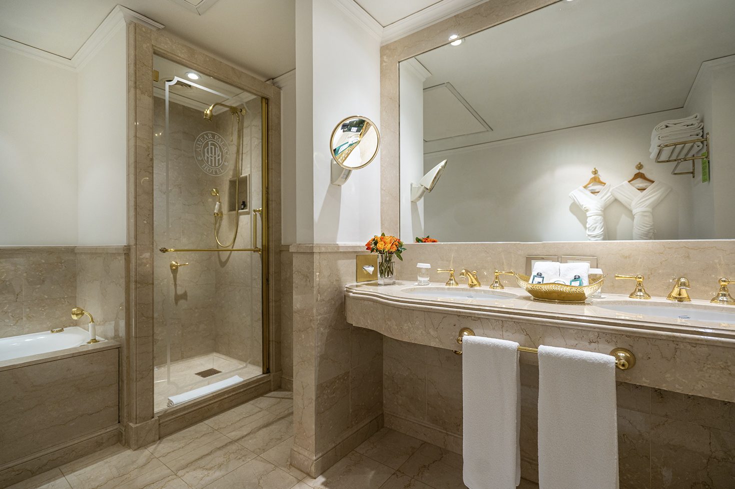 A luxury bathroom with a large mirror, shower and sink with brass finishes, complimenting the marble tiles.