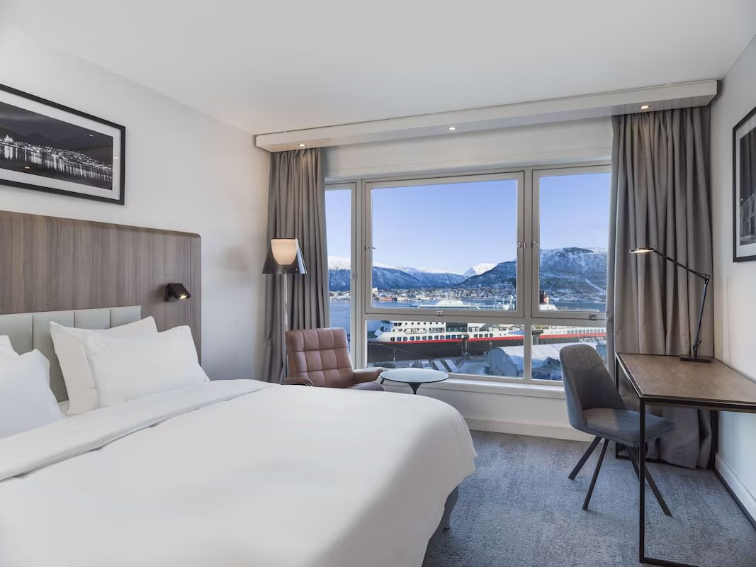 A bright bedroom in the hotel featuring a large bed with pristine white bedsheets, a desk and a large window, showcasing the incredible views on Tromsø.