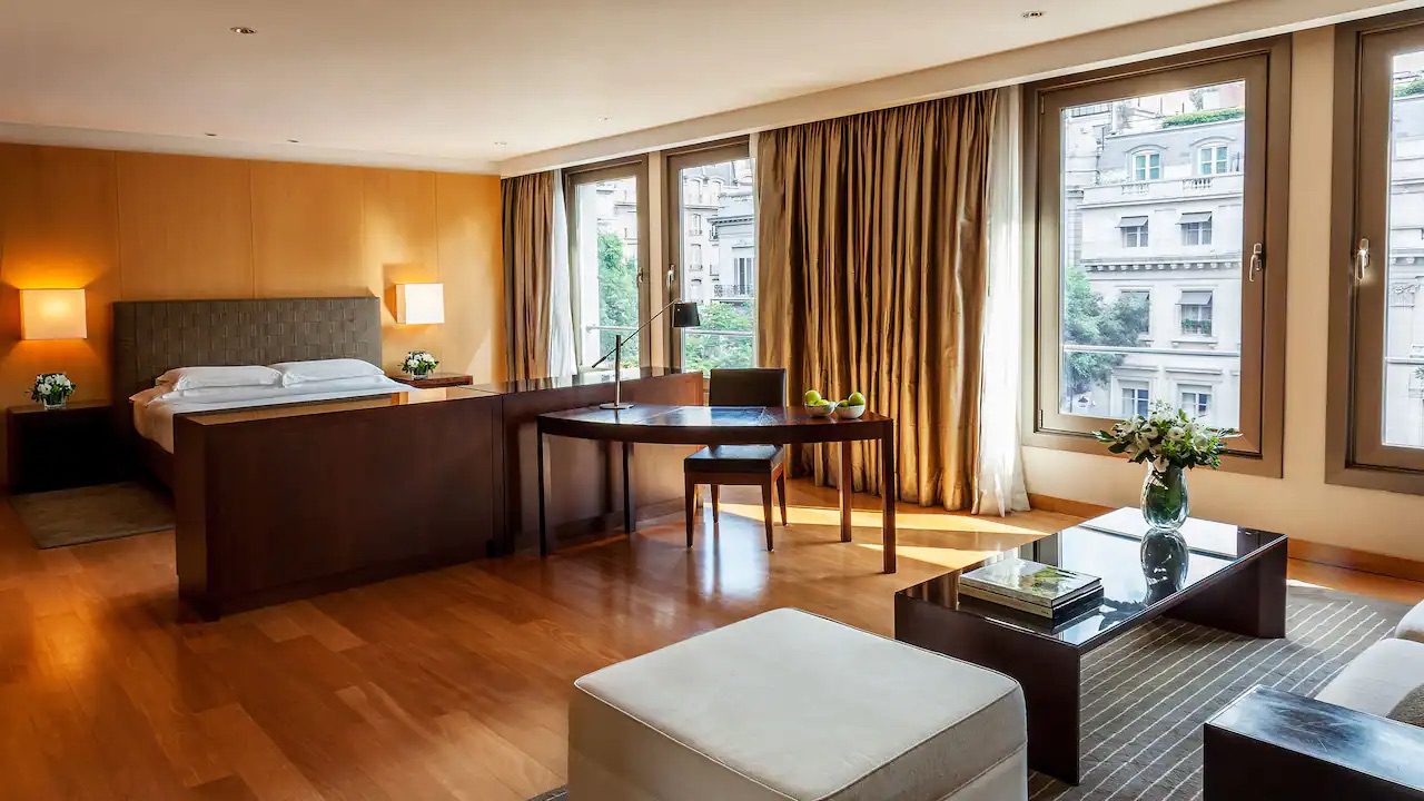 A spacious bedroom in the luxury hotel, showcasing a large bed, a desk, a sofa, coffee table and large windows showing the views of buenos Aires.