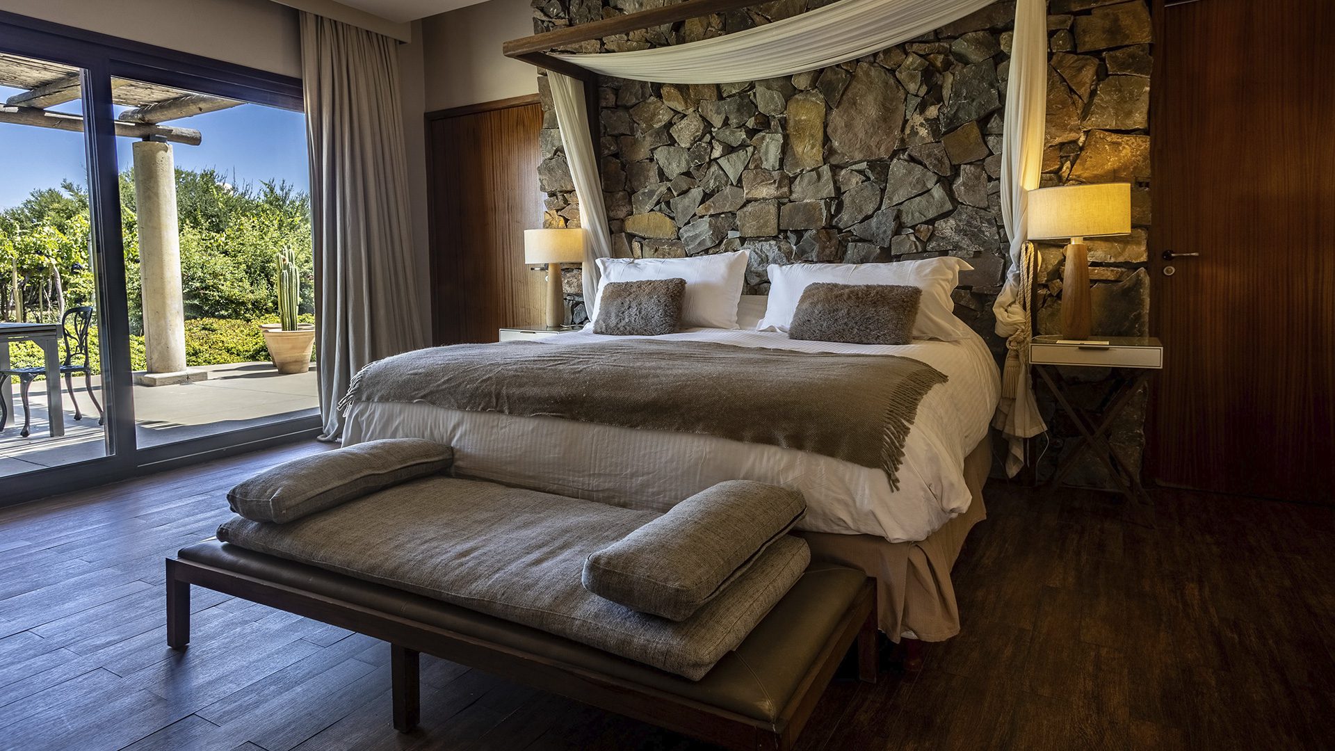 A luxury bedroom with a large bed, an exposed stone wall, wooden features and large glass doors opening onto the private garden.