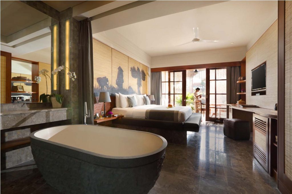 A view of the bedroom from the bathroom, with a luxurious bathtub in the foreground. In the background shows a large bed, a wooden desk, a large flatscreen tv and with a private balcony complete with chairs and tables facing out onto an incredible view.