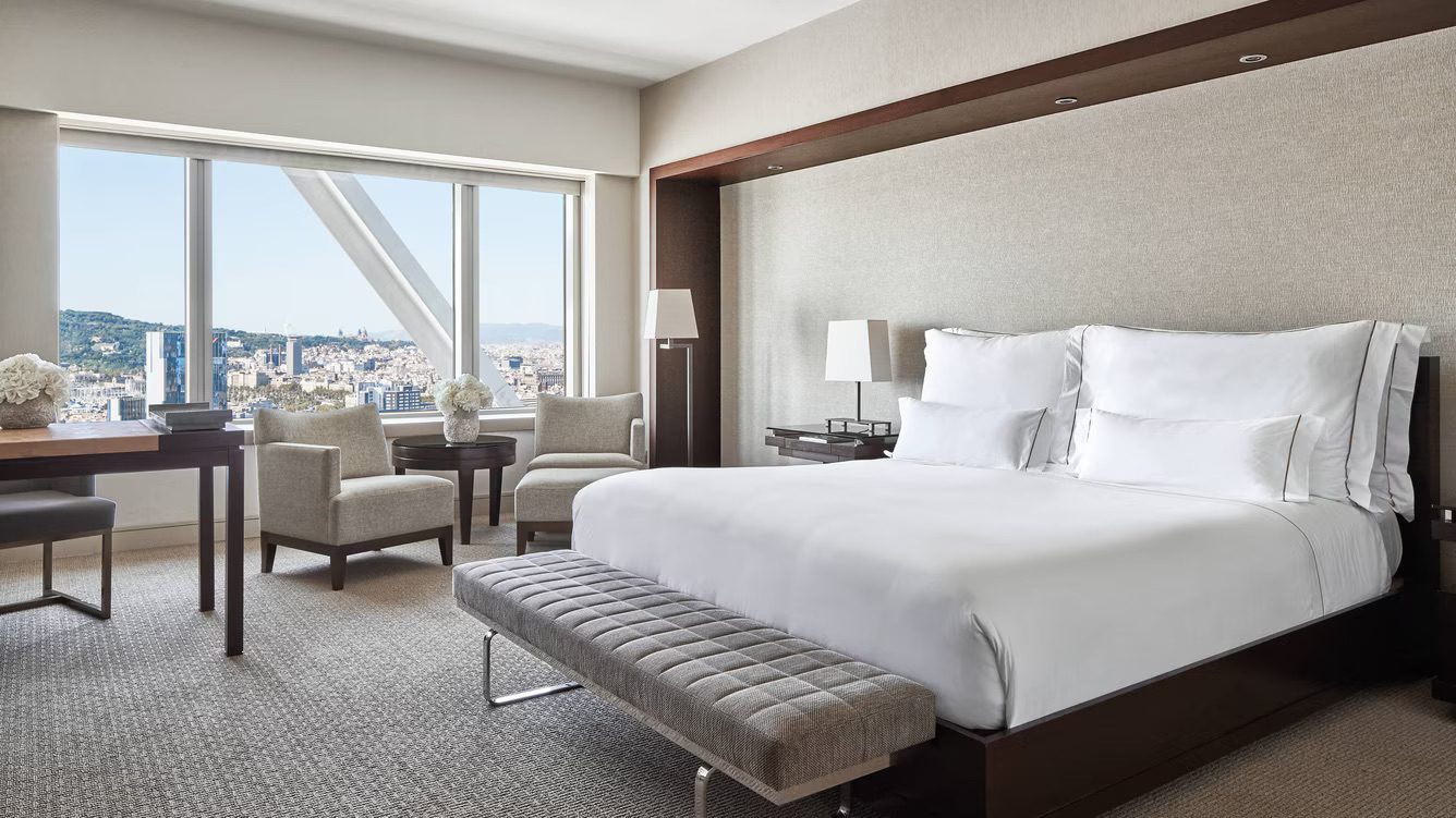 A spacious bedroom at the luxury hotel featuring a large bed with pristine white bedsheets, two armchairs, a desk and an incredible view of Barcelona.