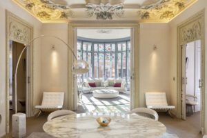 A suite in the luxury hotel featuring intricate architecture on the ceiling and around the door frames. A table and chairs in the foreground and a sofa in front of a stained glass window in the background.