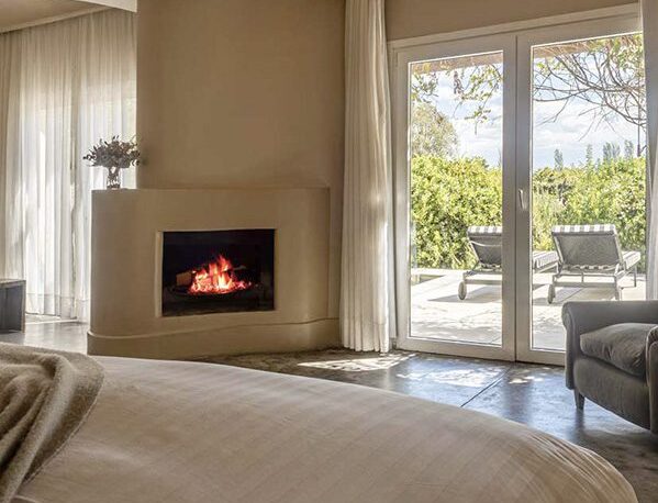 A luxury bedroom with a bed, chair, a log burning fire and doors opening up onto the private patio.