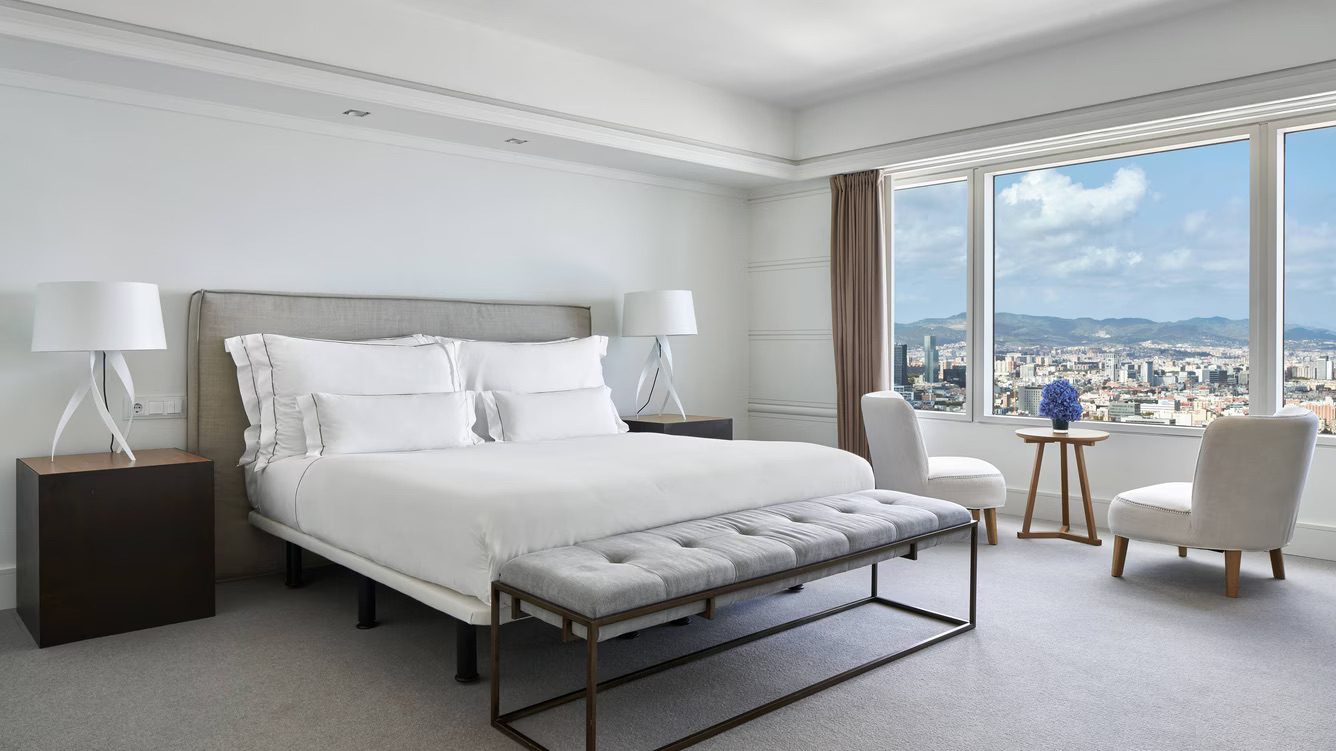 A spacious bedroom at the luxury hotel featuring a large bed with pristine white bedsheets, two armchairs and an incredible view of Barcelona.