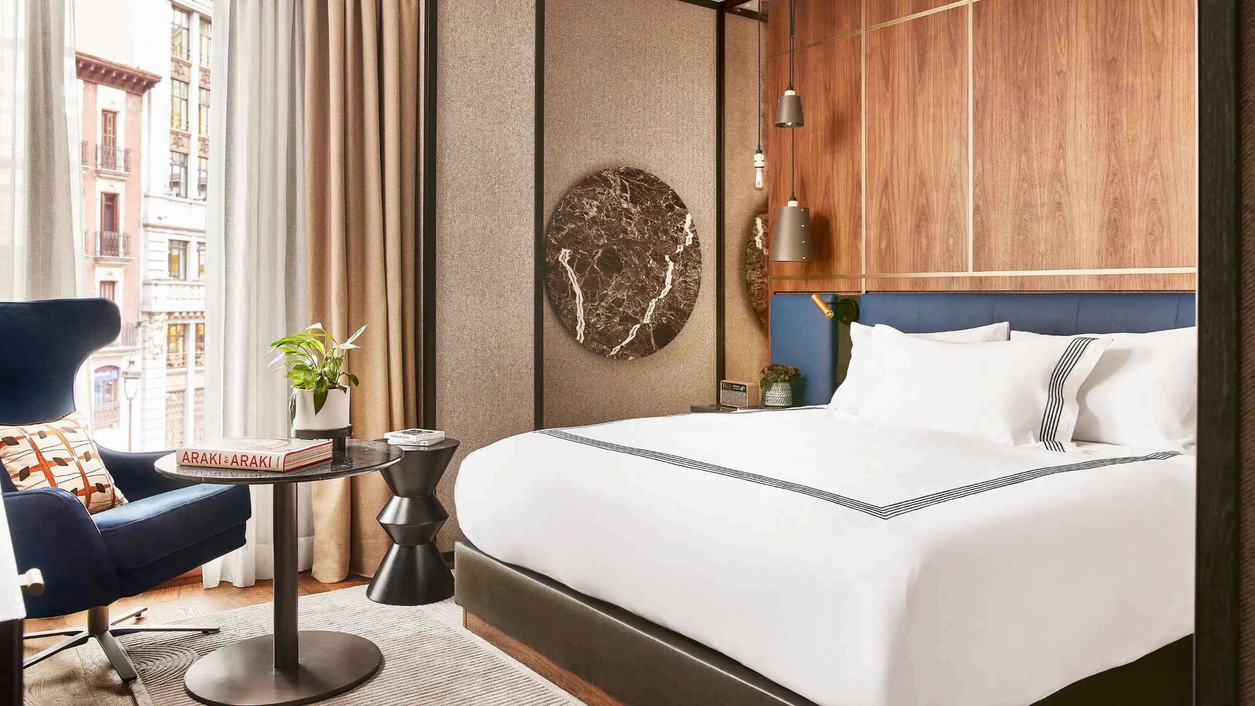 Luxury Bedroom at the Thomson Hotel in Madrid with a double bed with white linen, a leather armchair and coffee table with a window.