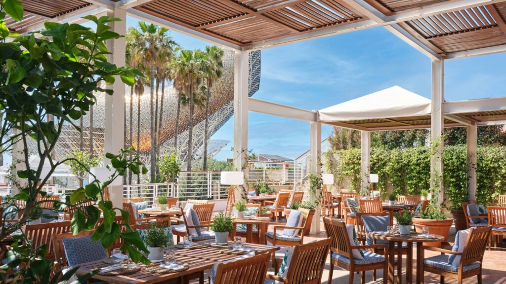 A spacious outdoor dining area at the luxury hotel, with canopies covering the tables and chairs, greenery and views of Barcelona.