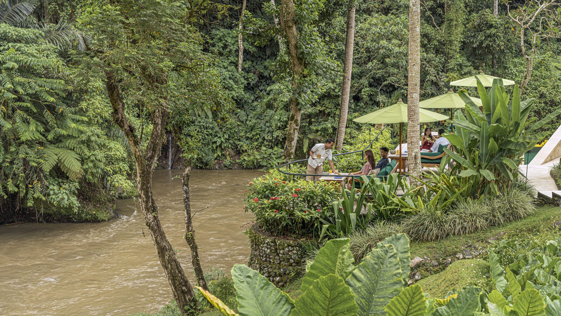 People eating in an outdoor dining area along a Balinese river, surrounded by incredible trees and greenery.