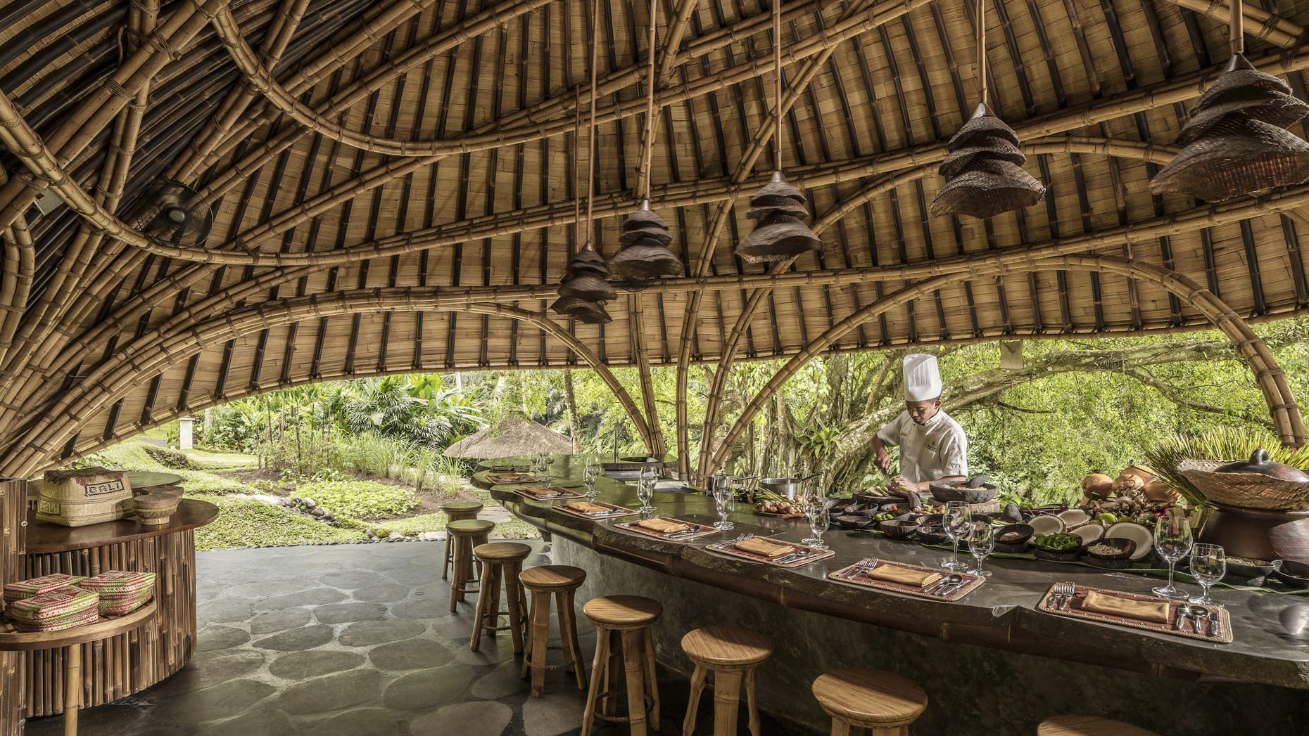 A chef preparing food for a secluded dining experience at Sokasi's riverside kitchen with a bamboo canopy structure and traditional Indonesian furnishings to complete the luxury experience. Stools and placemats surround the chef's kitchen so the guests can watch the food being prepared in real time.