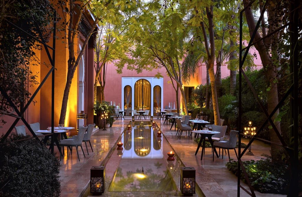 A serene courtyard with a water feature running down the centre and dining tables and chairs on either side and trees forming a canopy over the top.