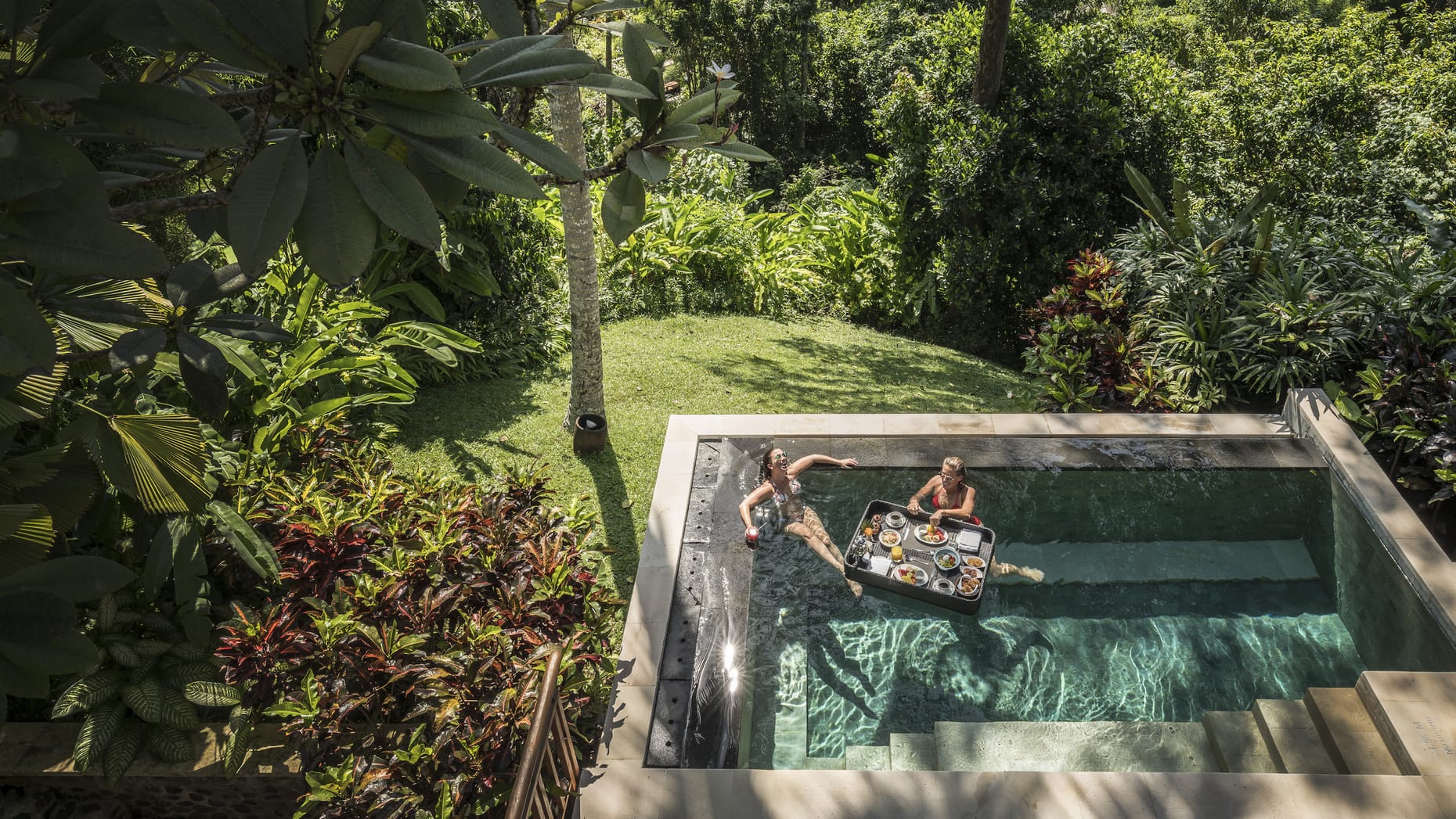 Two women enjoying a variety of breakfast foods floating on a tray in the secluded pool surrounded by lush greenery, perfect for your luxury vacation.