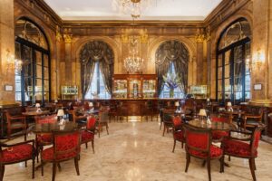 A luxury dining area with extravagant marble walls and ceilings, tables, chairs, a large bar the the back of the room, complete with large windows covered by elegant curtains.