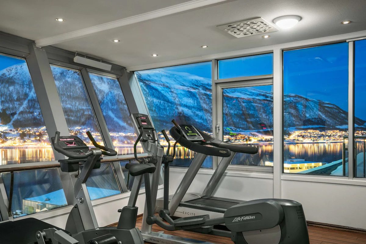 A gym with a variety of machines and windows surrounding the gym, showing the incredible mountain and lake views of Tromsø.