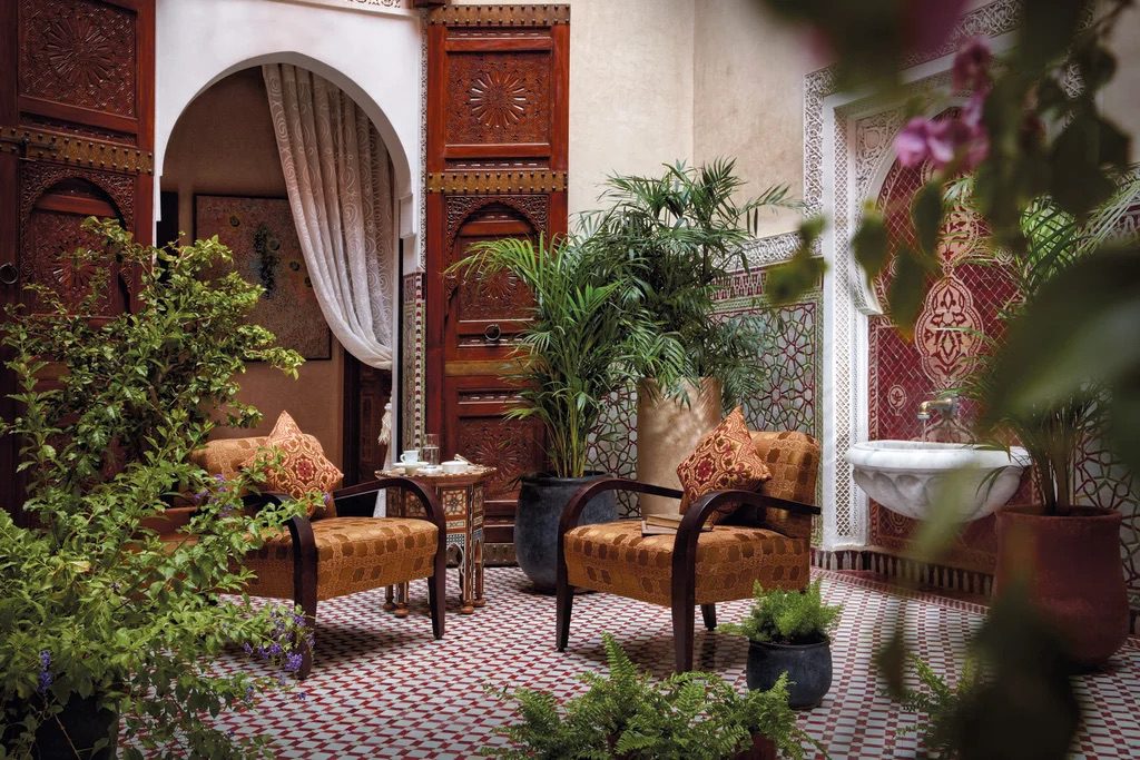 A luxury courtyard with two elegant chairs, large potted plants and intricate Morrocan designed doors, walls and flooring.