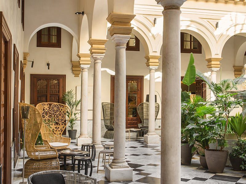 A luxury lobby with pillars and arches, a variety of potted plants, seating and tables.