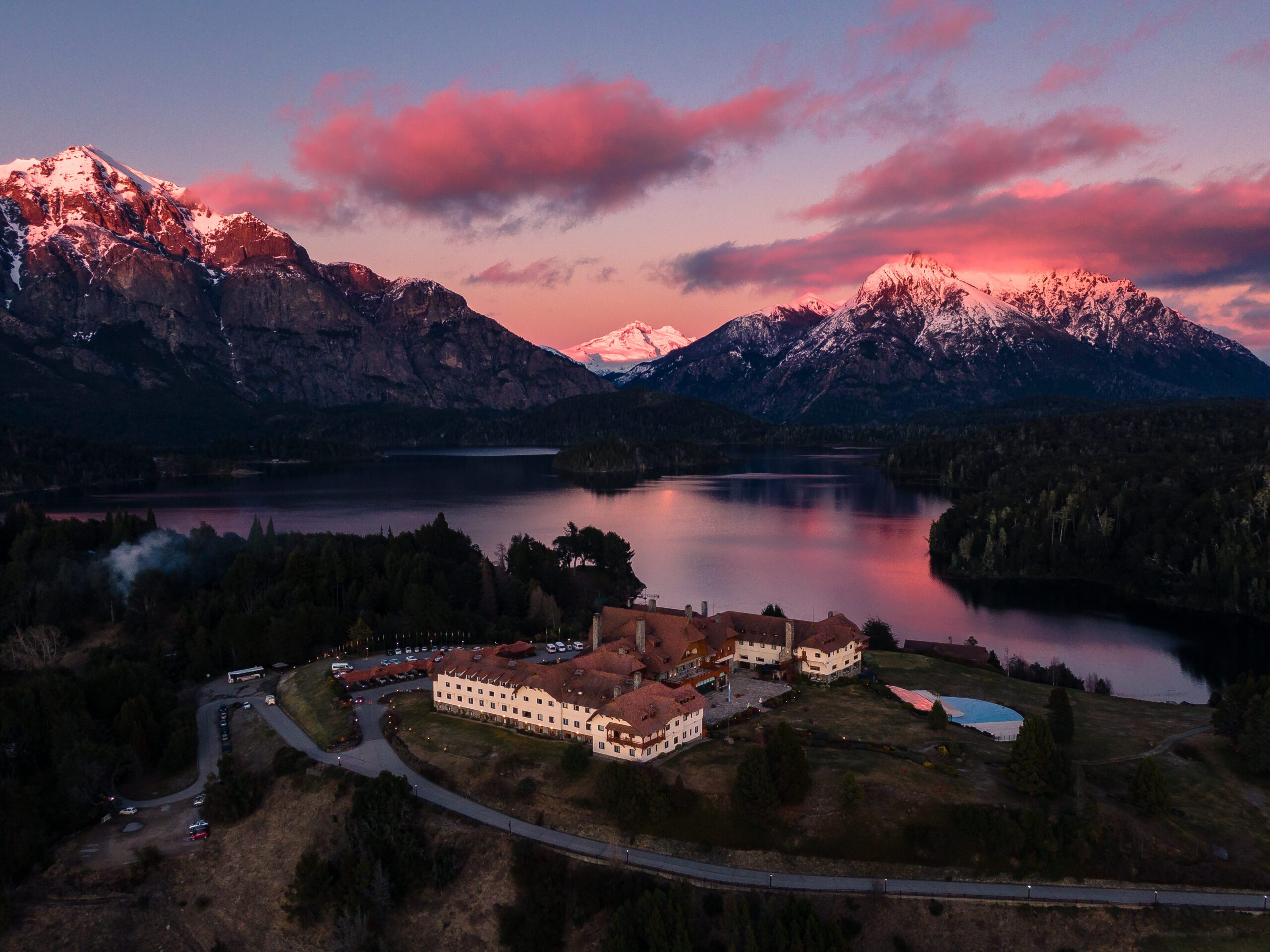 A view of the luxury hotel surrounded by greenery and incredible mountains behind, complete with a beautiful pink sunset.