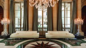 Two sofas in a corridor of the luxury hotel, large windows adorned with green curtains and pillars between them.