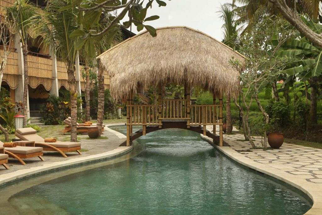 Luxury pool area with lounge chairs on the edge, in the centre is a thatched roof bridge that goes from one side of the pool to the other.