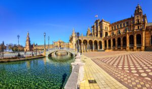 Plaza de España with the canal running up on the left under a small bridge and the main building which is red/orange in colour to the right and a pedestrian walkway in chequered red and cream tiles and mosaic on the right.