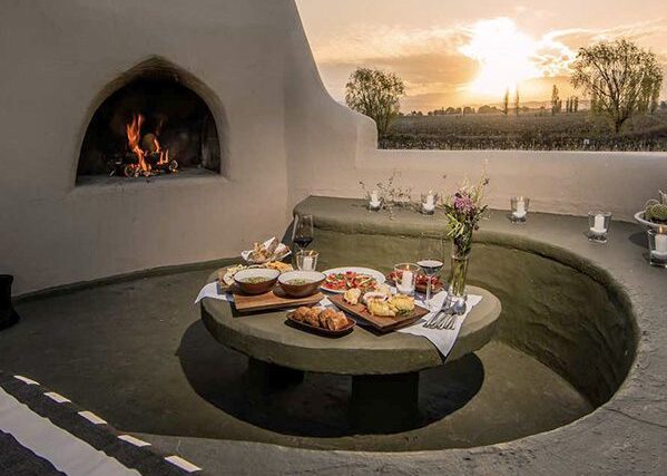 A private outdoor dining area with a log burning fire, a variety of food on a table in the centre and seating and candles around the edge.