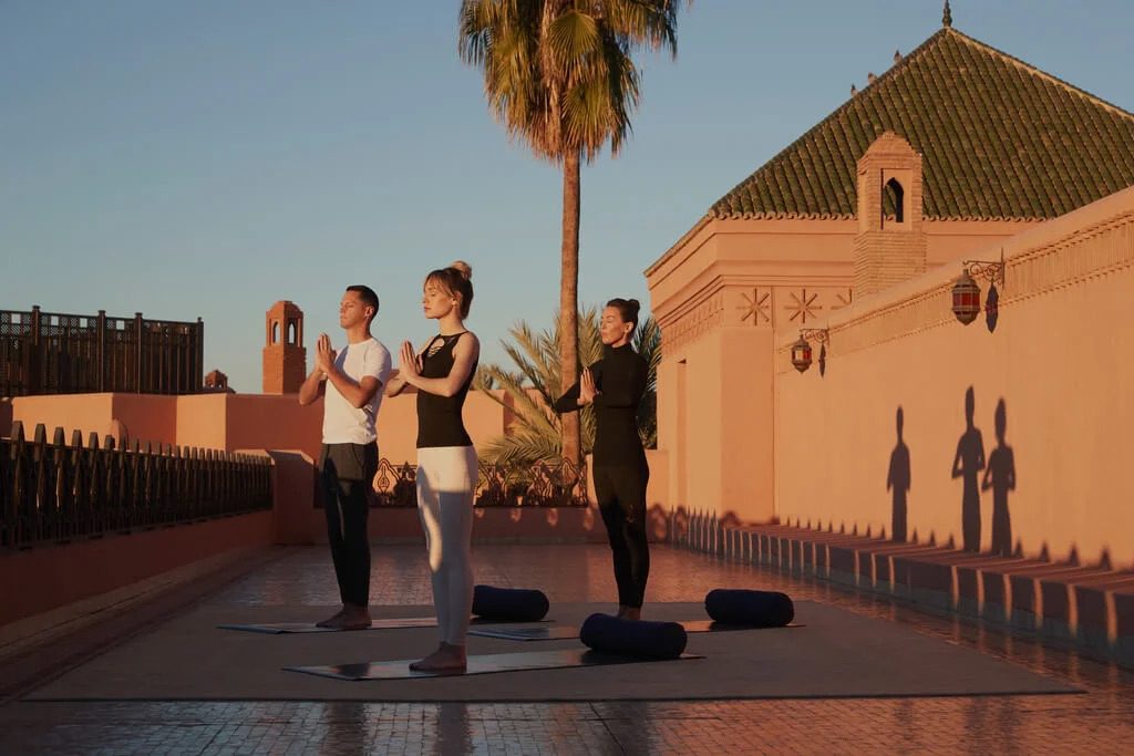 Three people enjoying yoga on a terrace at the Royal Mansour Hotel, with the incredible building and palm trees in the background.