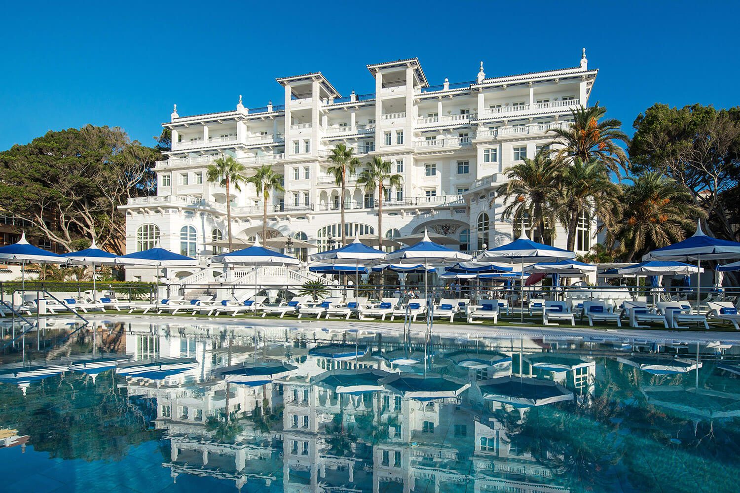 A view of the expansive outdoor pool with an array of sun loungers and umbrellas lining the pool and a view of the luxury hotel in the background.