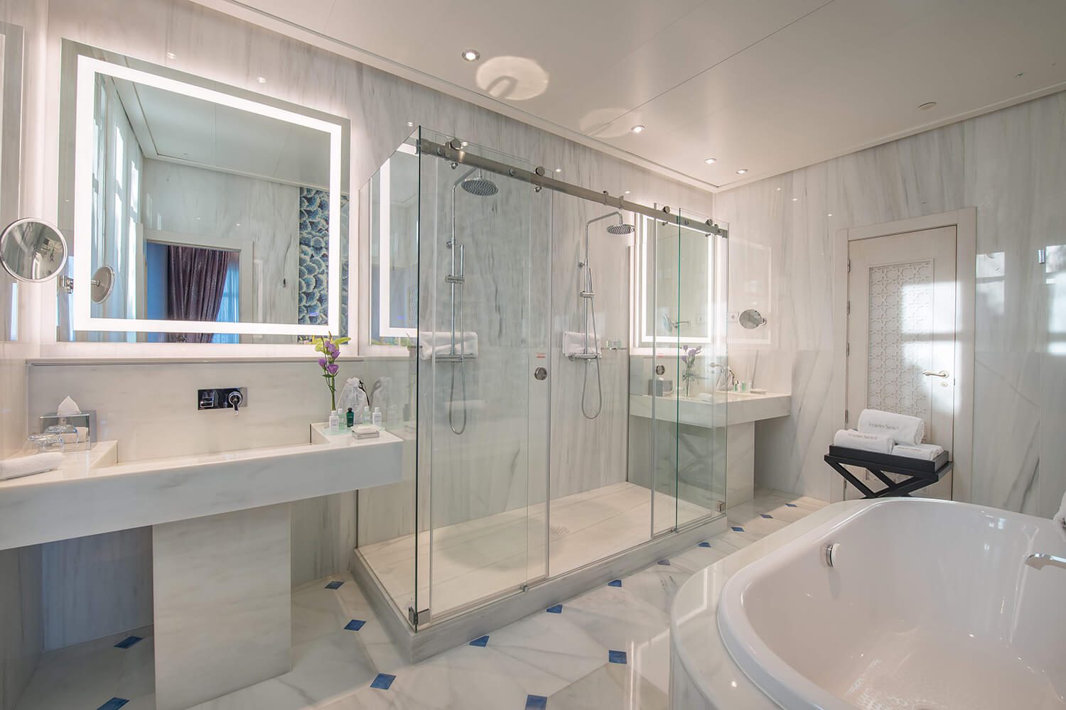 A bathroom at the luxury hotel with a spacious bathtub, a double shower and two sinks with lit mirrors above them.