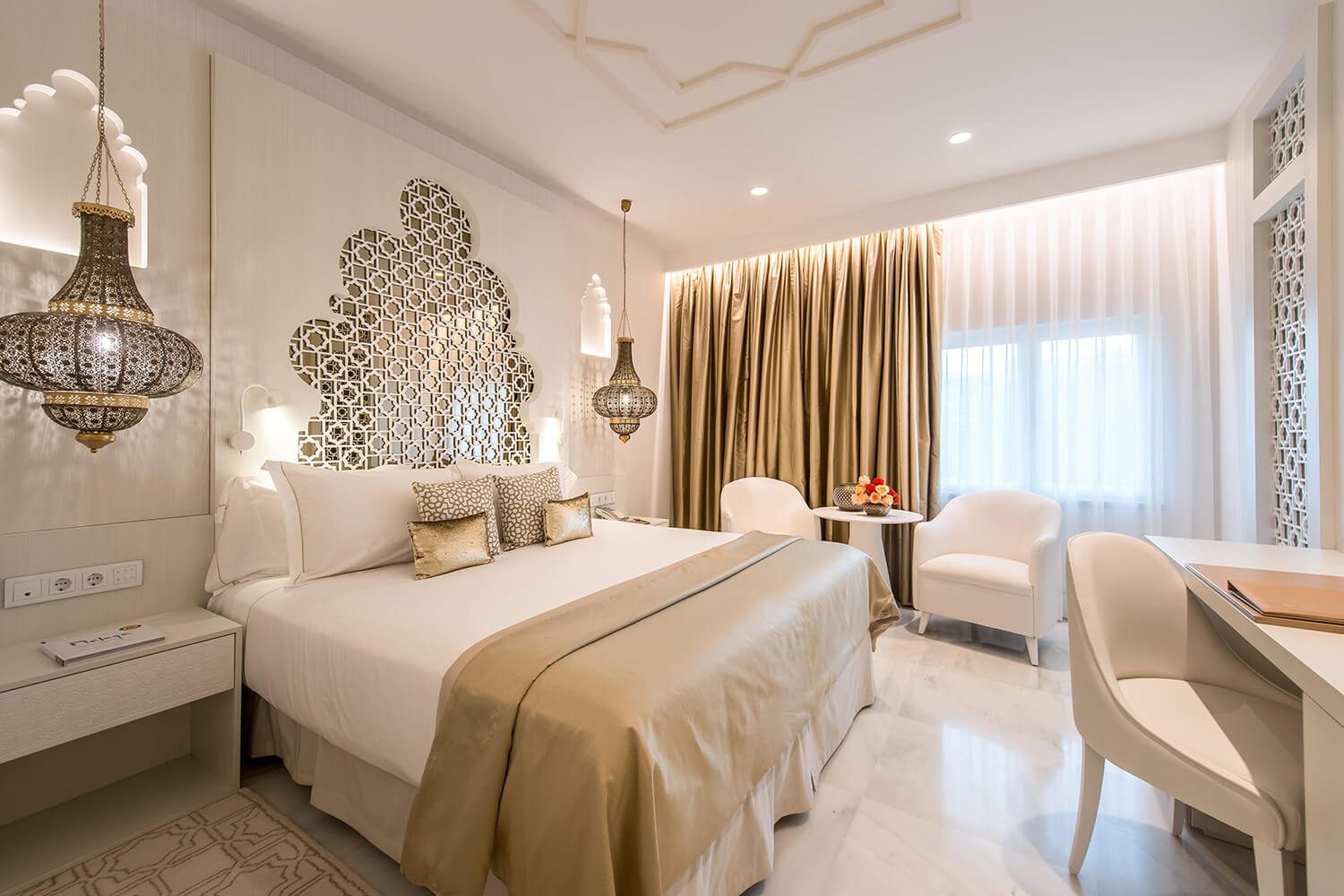 A bedroom in the luxury hotel, featuring a large bed adorned with white bedsheets, a desk and two armchairs next to a coffee table, finished with two hanging lamps either side of the bed.