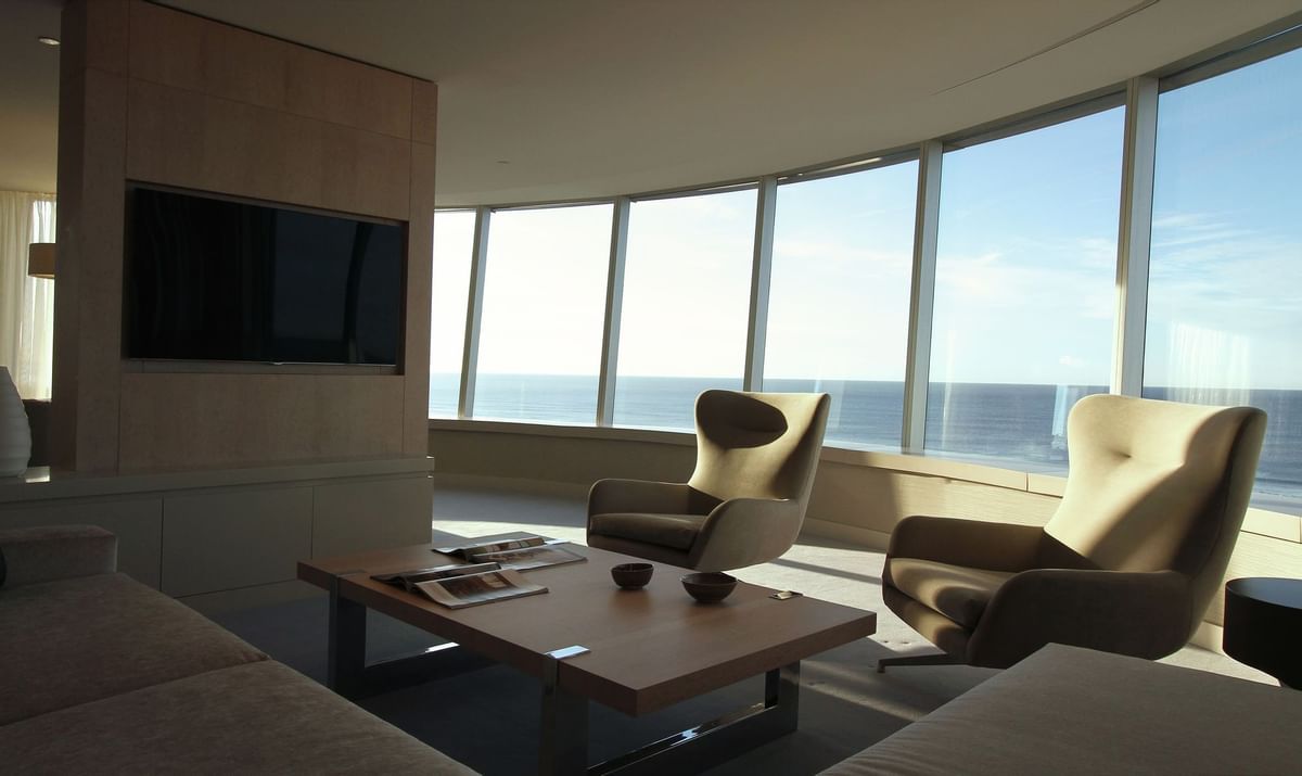 A living room, equipped with a large sofa, two arm chairs, a coffee table in the centre and a wall-mounted TV, complete with panoramic views of the ocean.