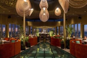 A stylish lounge with orange booth-style seating, bar, potted plants and brass pillars and architecture covering the walls and ceiling.