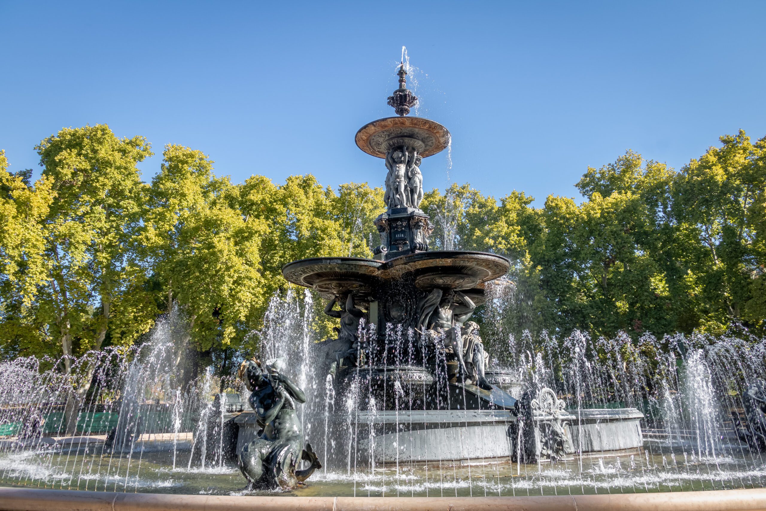 A fountain at General San Martin Park with intricate statues on the fountain, bright green trees and a blue sky behind.