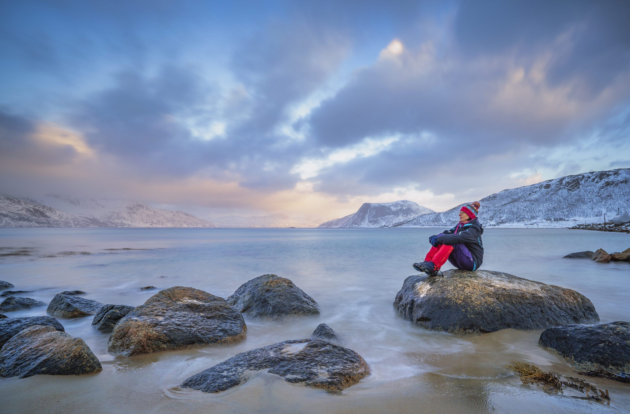 Woman sat on a rock looking out over the water and dressed in red and blue at polar dawn on Kvaløya Island and fjord near Tromso, northern Norway