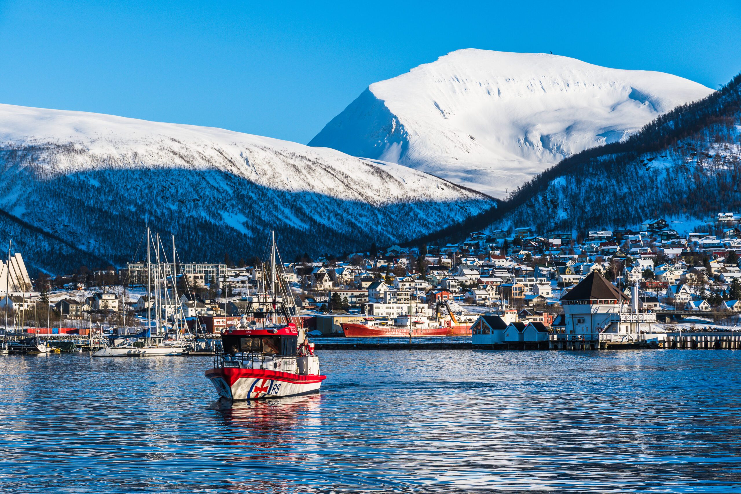 Tromso, Norway, View to the Tromso urban neighborhood Tromsdalen, in the foreground a boat from SAR Norwegian rescue service with snowcapped mountains behind under blue sky