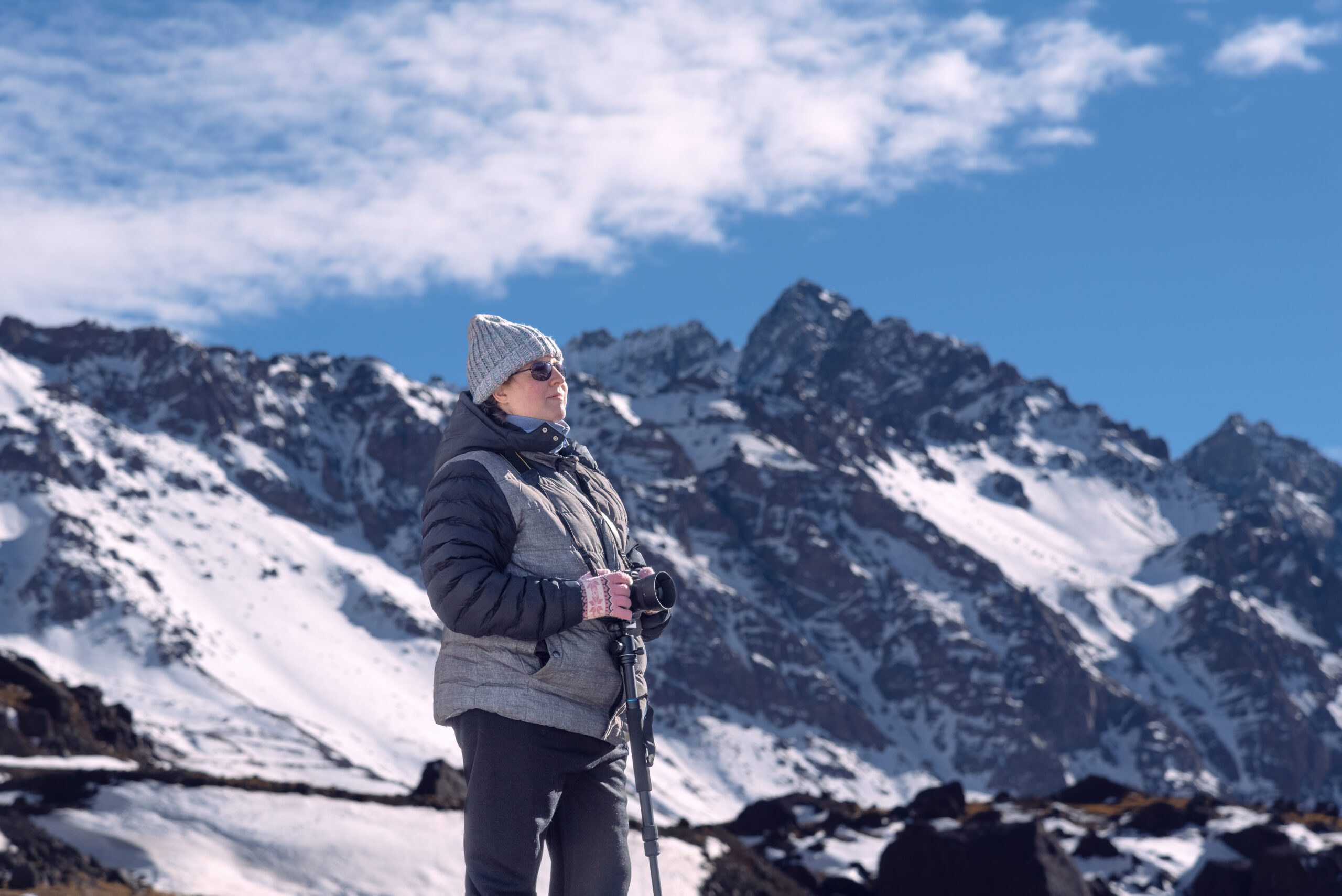 Solo Female Traveller on a luxury winter vacation with professional camera on the Mountain at the Bridge of the Inca in Mendoza Arentina. Woman is wearing winter clothing and hat and sunglasses with the mountain tops behind her and blue sky with white fluffy clouds