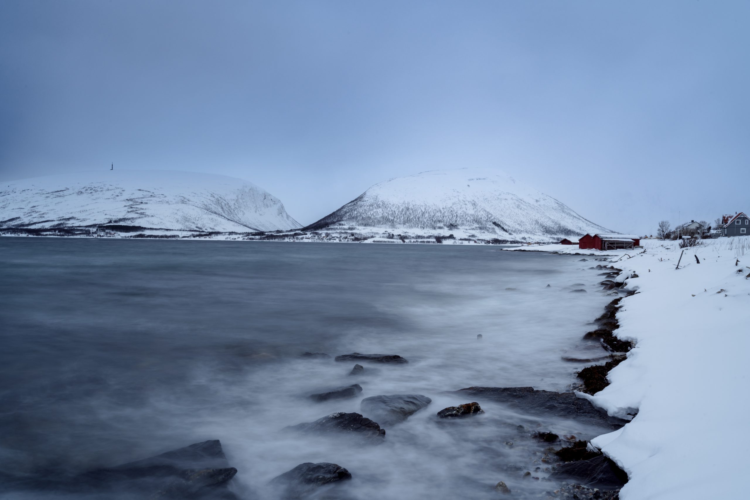 View of the coastline of Tromso in Norway showing snow and ice with snowvapped mountains in the distance and dark blue sky