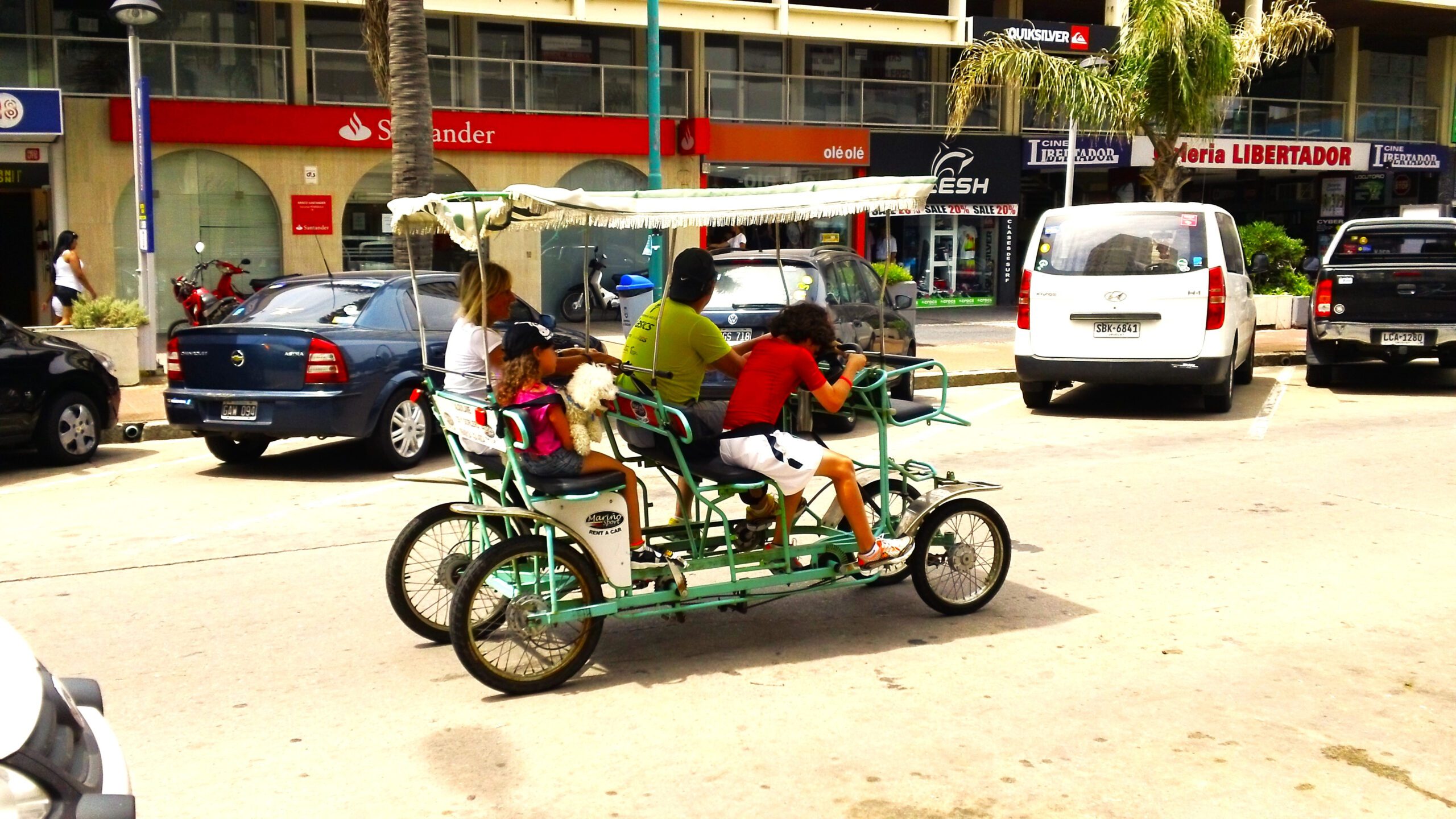 Four people riding a manual pedal quad bike with a sunroof and one person holding a small white dog. Travelling down the city roads in Punta del Este Uruguay.