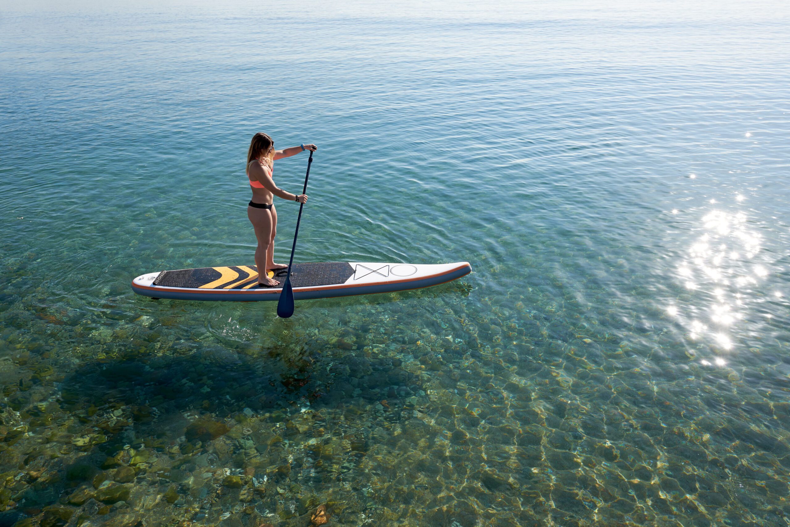 Side view of woman standing on a surfs up paddle board on clear and calm water with the sun glistening on the water