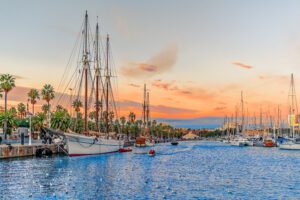 Evening seascape at Port Vell with a pirate galleon and many yachts against the backdrop of an orange-pink-blue sky in Barcelona, Spain.