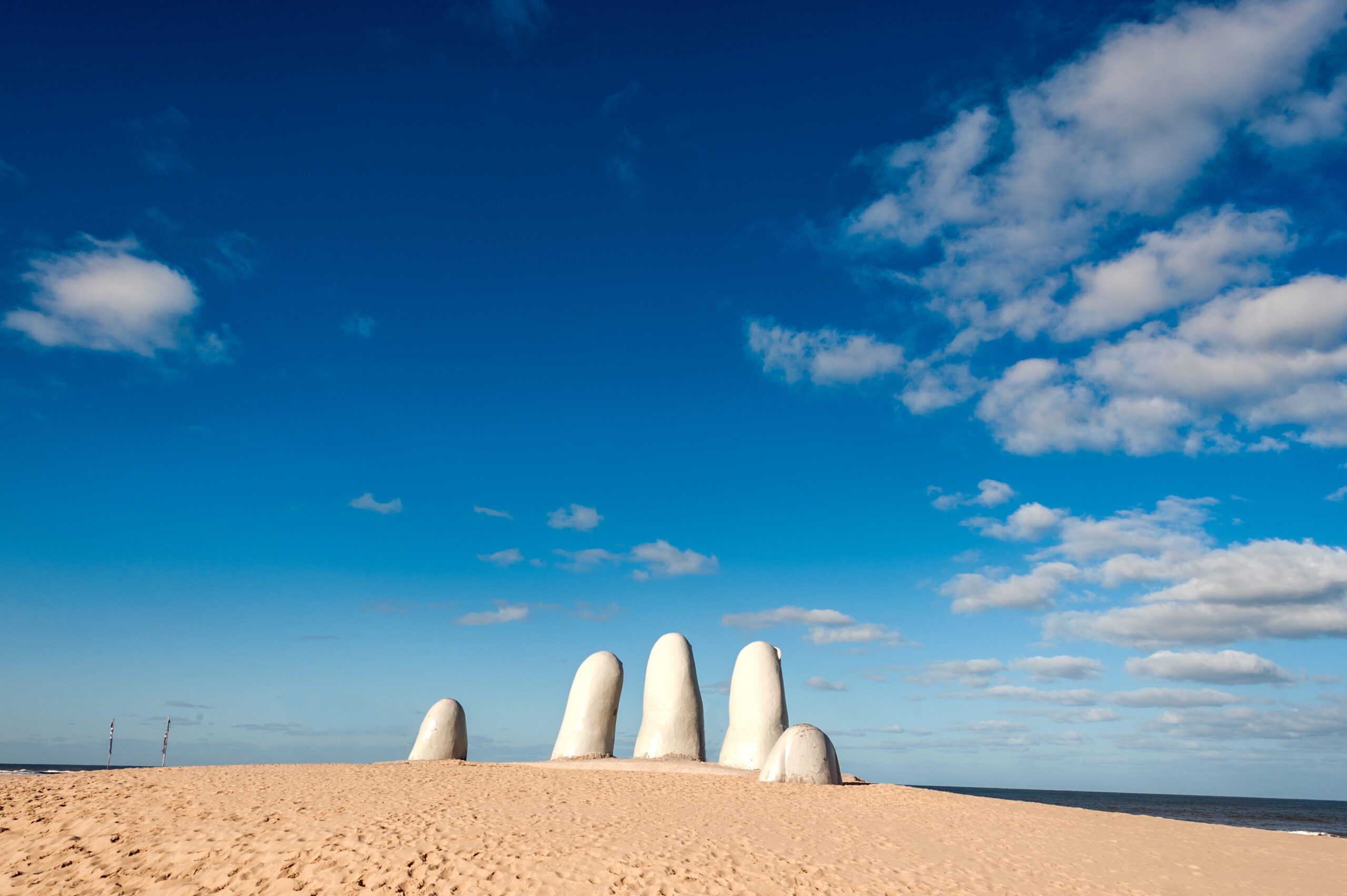 The fingers of the Hand of Punta del Este poking out the the yellow sand with blue sky above and a few white clouds.